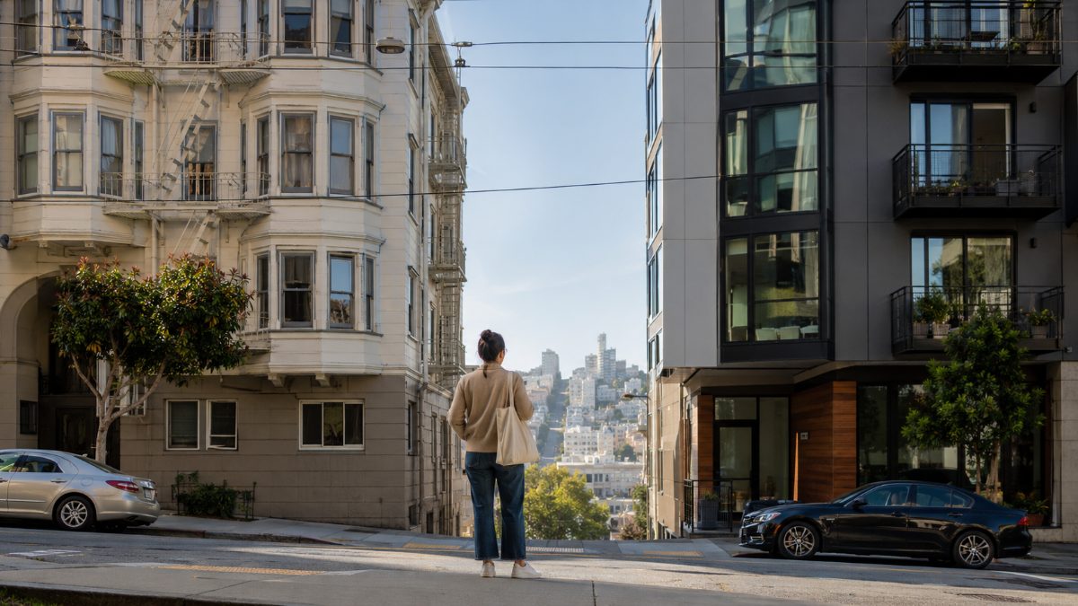 A person on a San Francisco sidewalk between an apartment building and a condo building, weighing the rent-versus-buy decision as San Francisco rent nears $4,100.