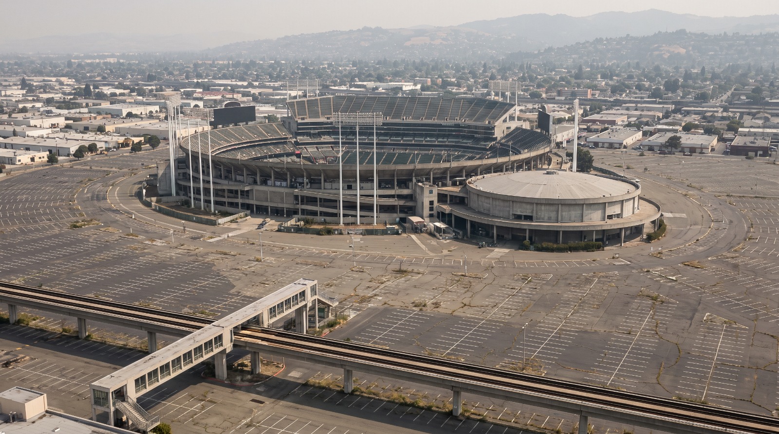 The Oakland Coliseum complex and its empty parking lots seen from above, with the BART guideway and the East Oakland flatlands behind it.