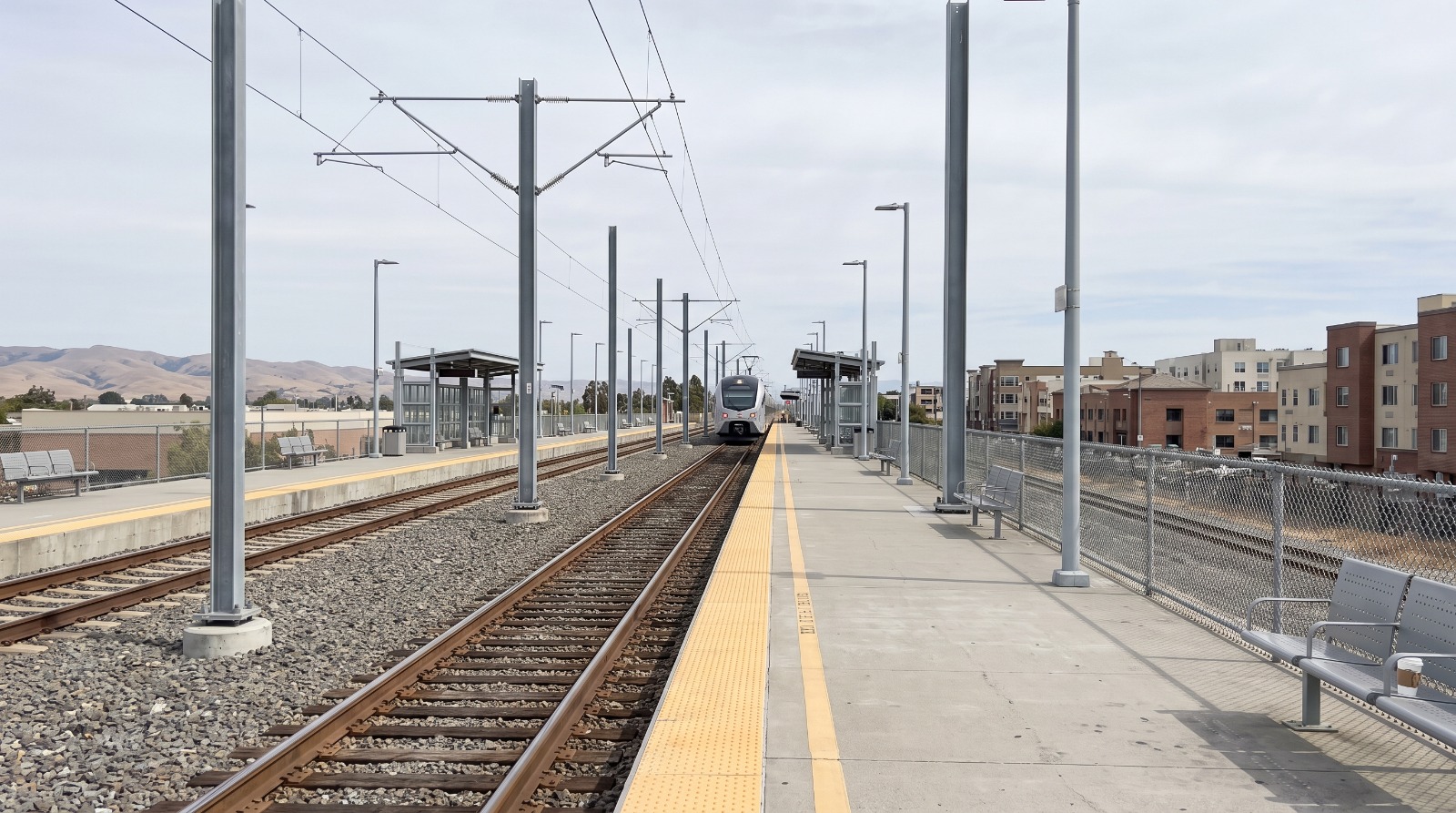 The Redwood City Caltrain platform with new electrification catenary wires and the low-rise downtown skyline behind it