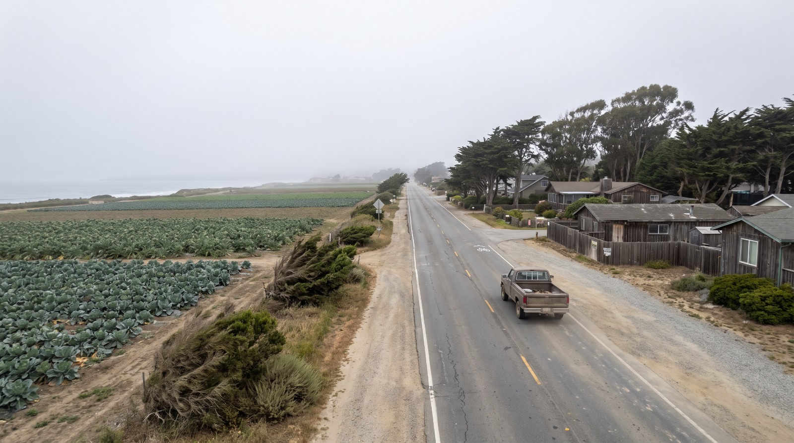 Kelly Avenue in Half Moon Bay looking west toward the Pacific under coastal fog, with farm fields and low coastside buildings.
