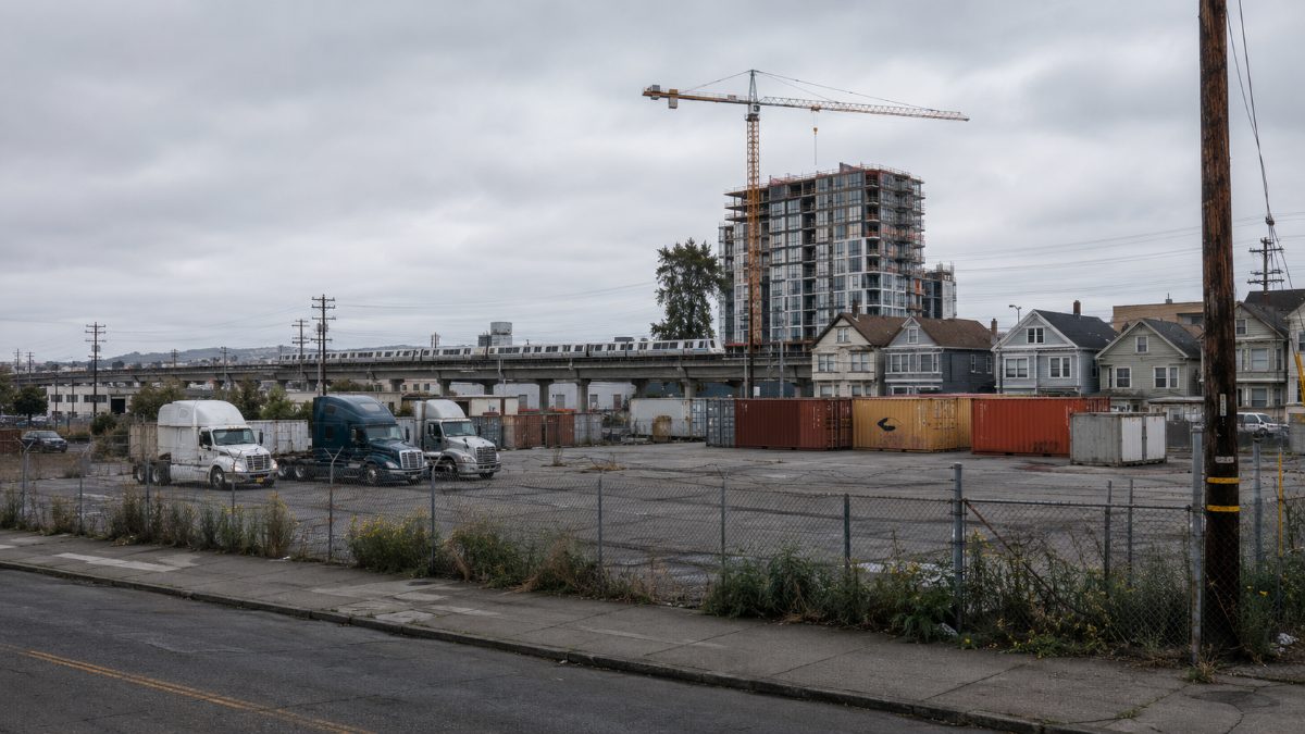 A fenced West Oakland lot holding parked trucks and shipping containers across from elevated BART tracks, illustrating stalled West Oakland development.