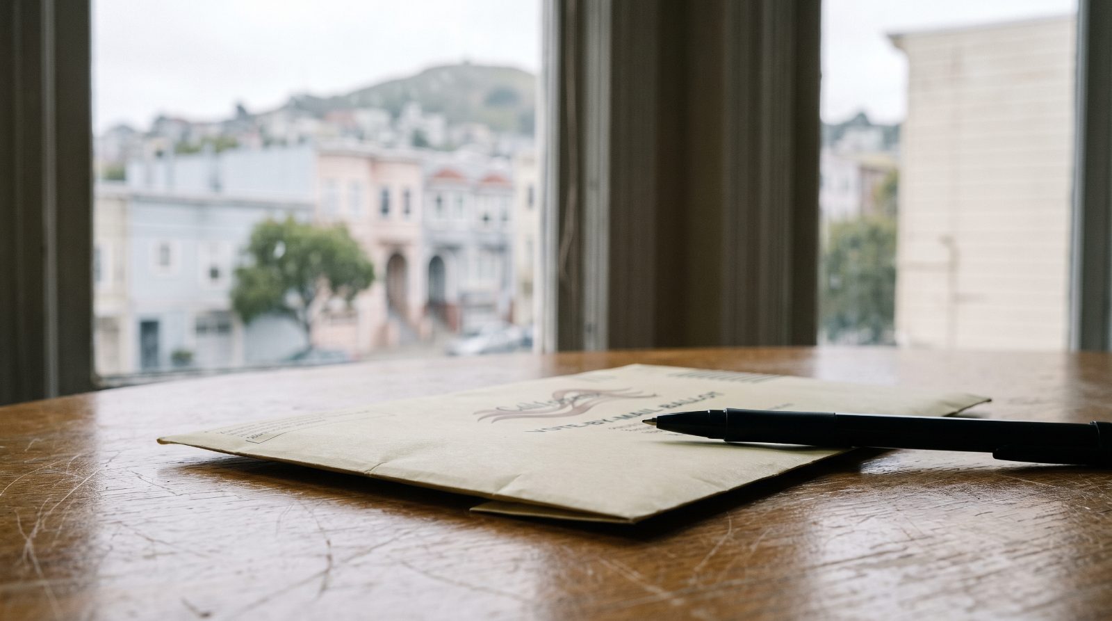 Vote by mail ballot envelope and pen on a kitchen table with Bay Area row houses outside the window