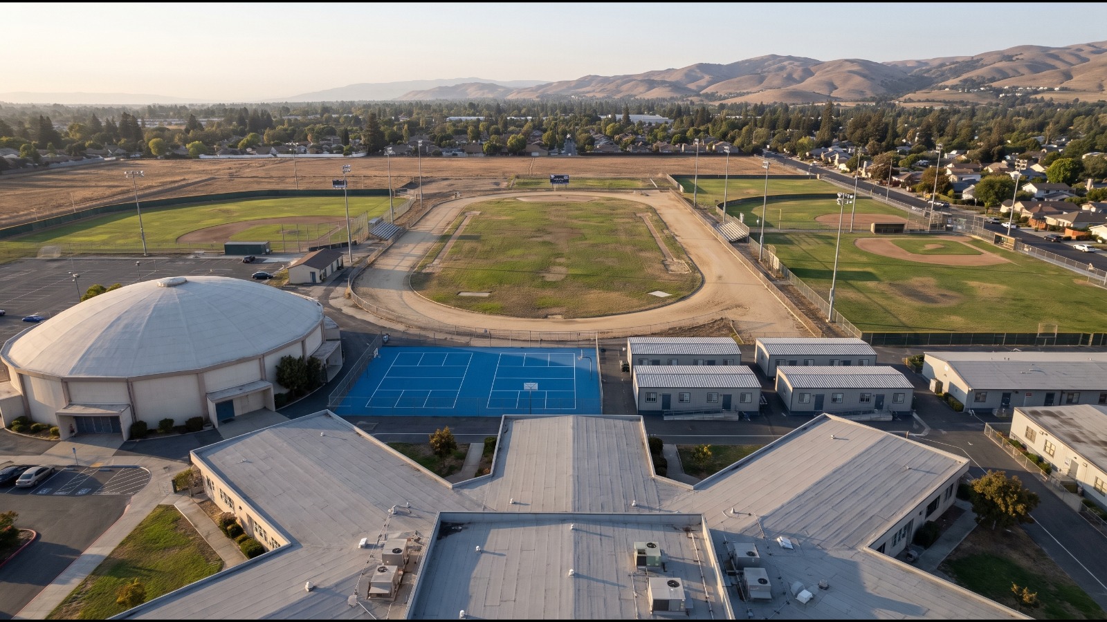 AI-rendered oblique aerial view of American High School in Fremont, its worn track and field ringed by baseball diamonds and homes, with the East Bay hills behind