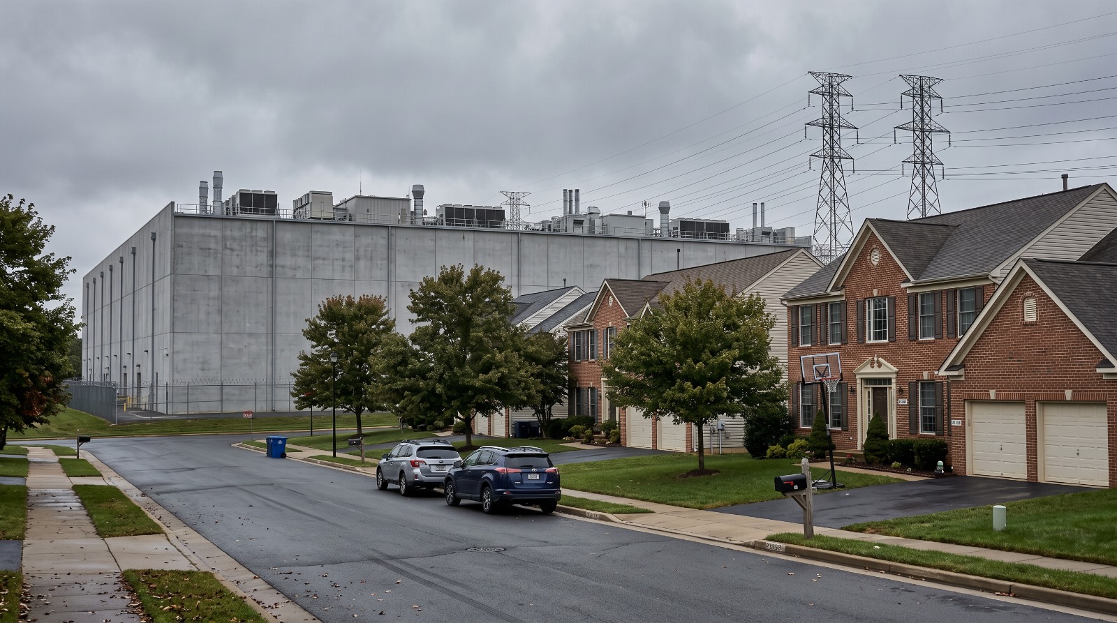 Suburban houses in Ashburn, Virginia with a data center building and high-voltage transmission towers rising directly behind them, the setting for the question of whether data centers hurt home values.